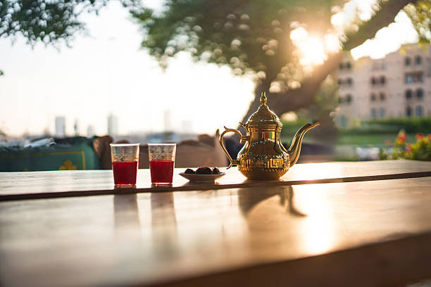 A serene outdoor cafe in Dubai features a traditional tea set with two glasses, sunlight streaming through lush trees, and soft shadows on a wooden table. The scene exudes warmth and tranquility during sunset.