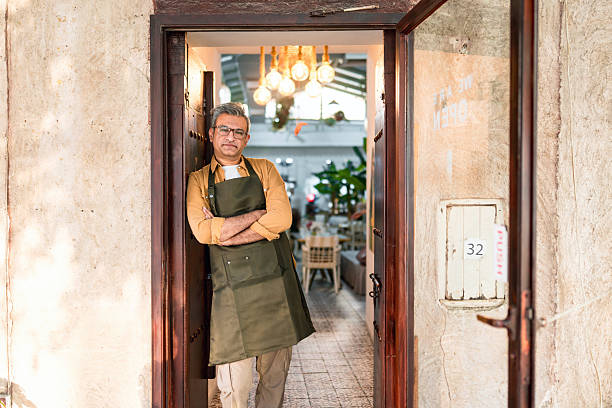 Middle-aged Middle Eastern man, wearing a green apron over a mustard shirt, stands confidently in the doorway of his rustic cafe. Warm lighting and greenery are visible inside, creating a welcoming and cozy atmosphere.