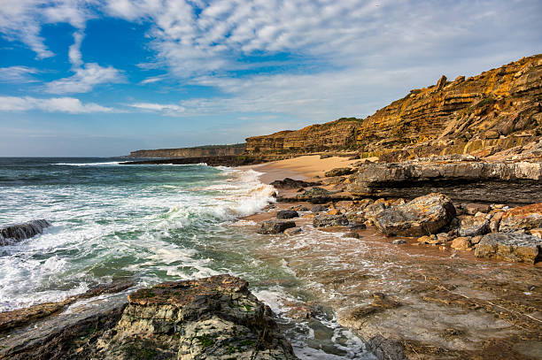 Waves crash against the rocky shoreline of Pedra Branca in Ericeira, Portugal, with layered cliffs and turquoise water.