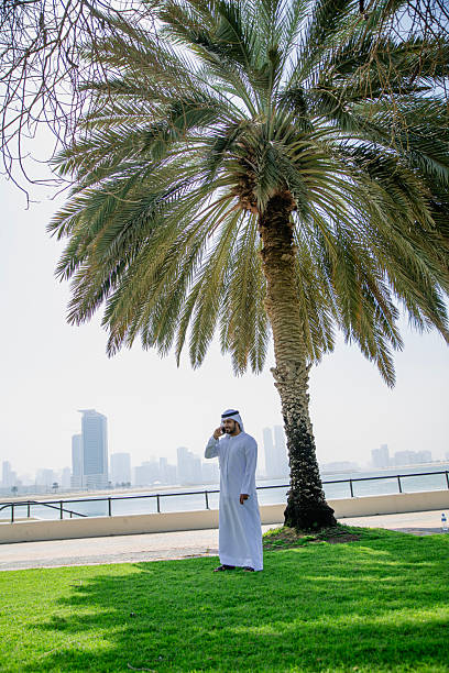 Portrait of middle eastern businessman with traditional emirati clothes, smiling, speaking with smartphone and looking away from the camera against Dubai cityscape, UAE