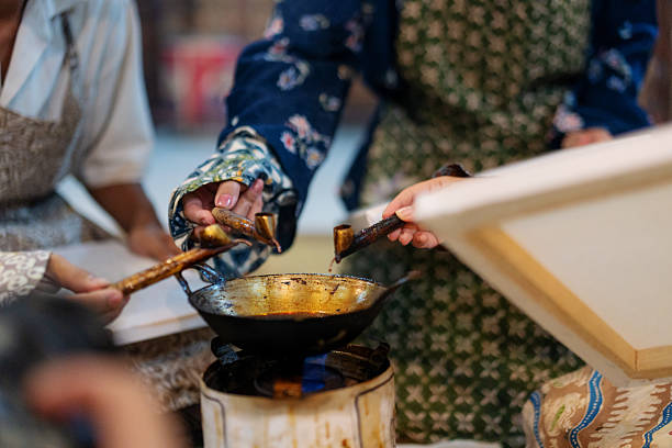 Top-down view of batik workshop participants dipping canting tools into melted wax during a traditional batik workshop in Indonesia. The scene captures the collaborative and cultural spirit of batik-making, featuring protective sleeves and hand-drawn floral patterns on fabric, ready for waxing.