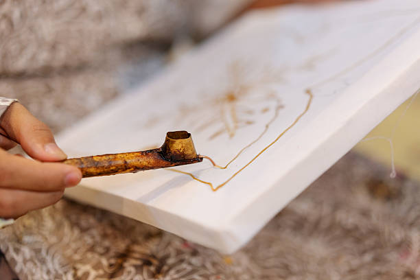 A close-up shot of a hand holding a canting tool filled with hot wax, used to draw intricate patterns on fabric during a traditional Indonesian batik-making process. The floral pattern emerges slowly as the wax seeps into the cotton canvas, capturing the artistry and cultural significance of batik craftsmanship.