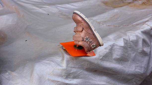 A small pink toddler shoe with flower decorations is mounted upside down on an orange base over a gray tarp.