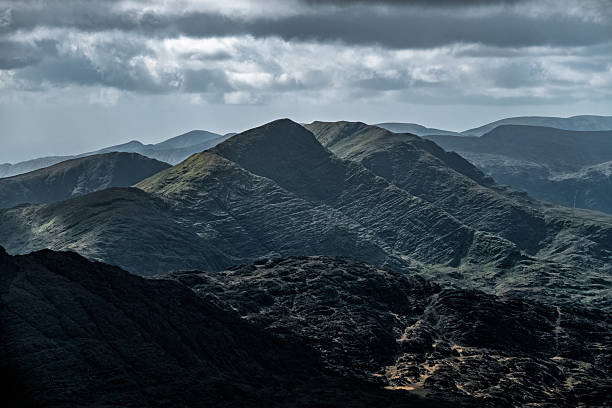 istock Dark, rolling hills under a cloudy sky. Shadows accentuate the rugged texture of the mountain slopes and valleys. Distant peaks are visible. 2210014858