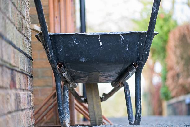 Close-up of an aged black wheelbarrow placed against a brick wall in an outdoor setting, symbolizing construction, gardening, and rustic charm