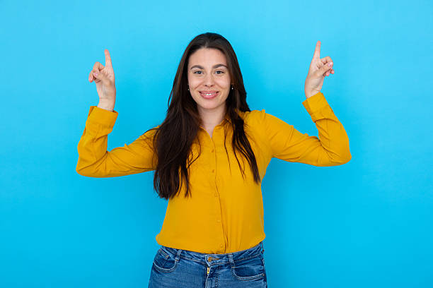 istock Woman pointing upward with index fingers, grinning while facing camera against blue backdrop 2210037171