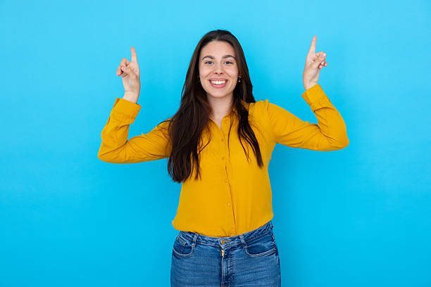 istock Cheerful female pointing upward with index fingers, beaming against blue backdrop 2210037175