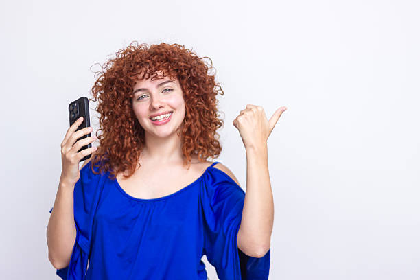Young woman showing her smartphone while pointing her thumb to the side and smiling at camera