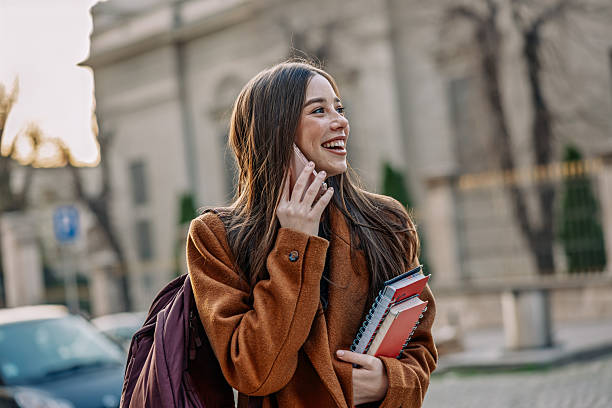 istock Smiling young woman walking outdoors and talking on the phone while holding notebooks 2210073965