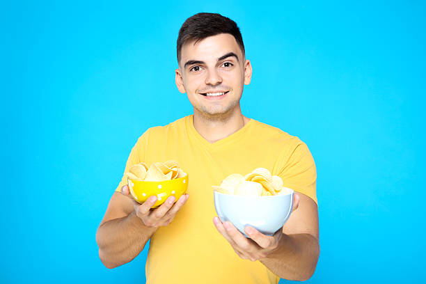 istock Young boy holding potato chips in bowls on blue background 2210084150