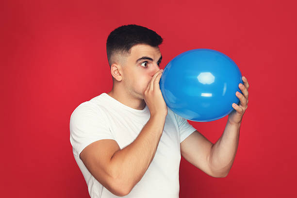 istock Yuong man blowing balloon on red background 2210085690
