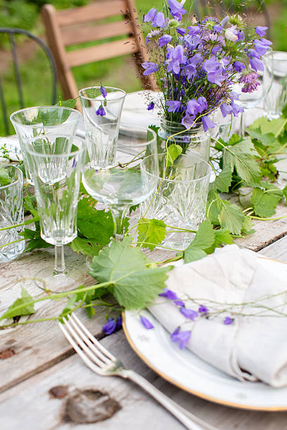 Detailed view of a rustic outdoor table set with crystal glasses, linen napkins, and purple wildflowers. Green leaves and natural elements create a charming Scandinavian summer vibe. Ideal for searches related to eco-friendly dining, table setting inspiration, or garden party decor.