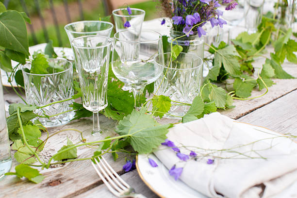 Detailed view of a rustic outdoor table set with crystal glasses, linen napkins, and purple wildflowers. Green leaves and natural elements create a charming Scandinavian summer vibe. Ideal for searches related to eco-friendly dining, table setting inspiration, or garden party decor.