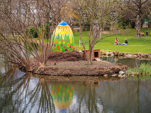 Innsbruck, Austria - March 21, 2025: Colorful Easter egg on a small island of Rapoldi pond, with trees and grass in the background. Two people are seated on the grass nearby