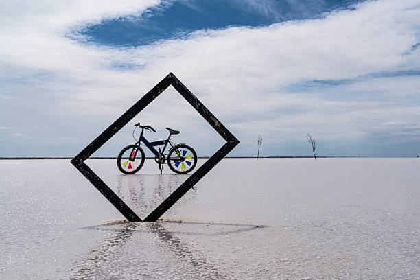 An image of a bicycle framed in a salt lake. The picture frame was dipped into salt from the corner, and the bicycle visible in the background was viewed through the frame. There is an impressive sky image. It was taken with a full-frame camera in daylight.