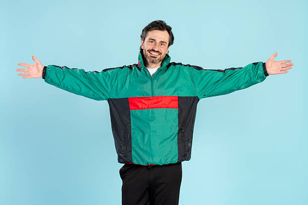 Smiling man with open arms, wearing a vibrant, colorful 90s-style jacket, poses against a blue background, radiating joy and a welcoming attitude with cheerful body language