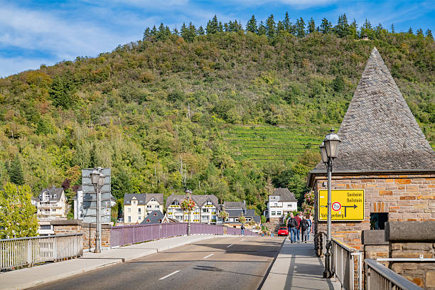 A bridge over a calm road with beautiful houses lined up along the street, with vineyards in the Moselle Valley, Cochem's scenic backdrop.