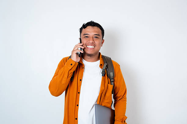 istock Image of handsome young Asian student wearing backpack and holding laptop using smartphone for phone on isolated white background 2210150526