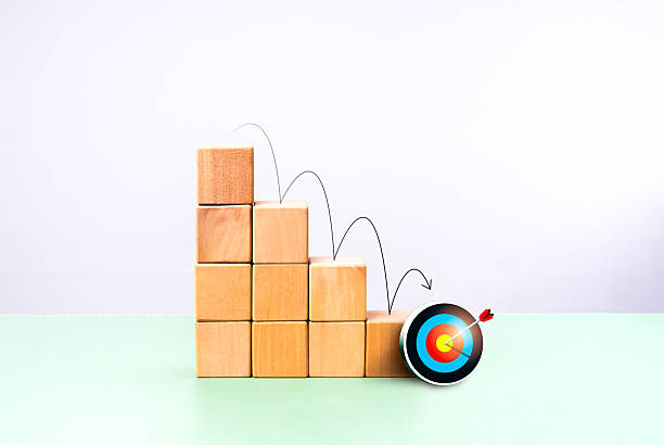istock 3d colorful target dart icon at the bottom of wooden cube blocks as bar graph chart steps with arrow jumping down, on green table and white background. Business goal, planning, back to basic concept. 2210153243