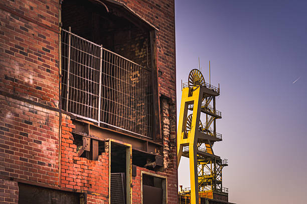 An old industrial building made of red brick, surrounded by a yellow conveyor tower that rises into the evening sky. The destroyed structures tell stories of past times, while an airplane strip on the horizon symbolizes modernity.