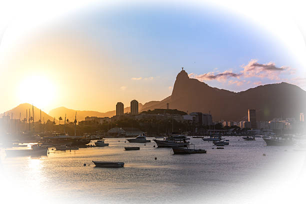 Photographed in Urca, Rio de Janeiro, this image captures the iconic silhouette of Corcovado Mountain overlooking the city during a golden sunset. Boats float in Guanabara Bay, and the buildings reflect the last rays of the day. The photo is edited in a soft, dreamy style with a white vignette that frames the composition.