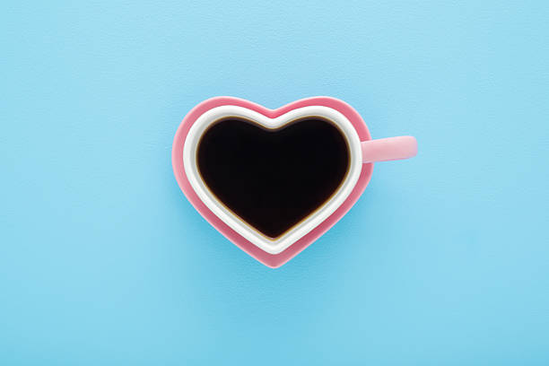 Heart shape white cup and pink saucer. Fresh black coffee cup on light blue table background. Pastel color. Closeup. Top down view.
