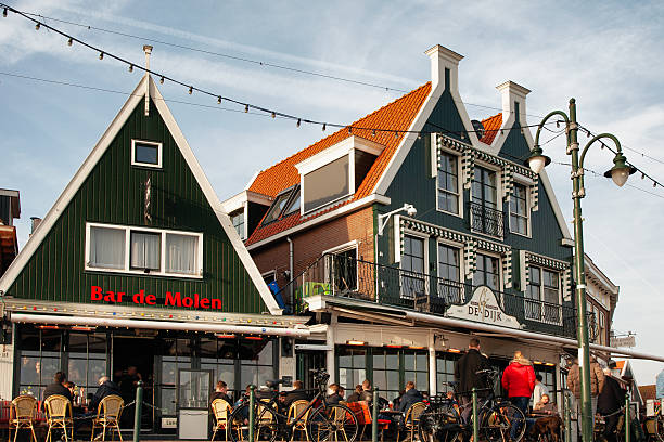 Volendam, Netherlands. February 16th, 2019: A bustling terrace outside Bar de Molen, with people enjoying the warm weather. The charming green and white buildings typical of the region's architecture stand in the background, while string lights hang overhead, adding to the cozy atmosphere.
