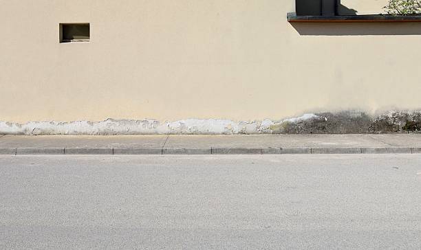 istock Yellow plaster facade peeled in the lower side for moisture problems. Concrete sidewalk and street in front 2210174620