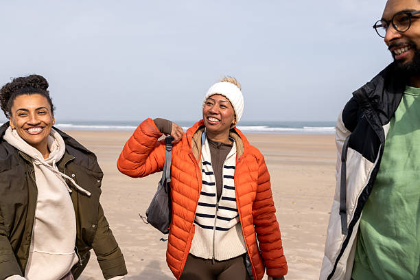 A wide shot of a diverse group of young adults enjoying each other's company on a cool beach day at Newton-by-the-Sea, Alnwick. All are warmly dressed, sharing a casual, joyful moment by the ocean. They are engaging in conversations with each other while smiling.\n\nVideos are available similar to this scenario.