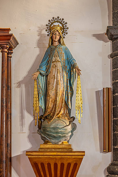 Parroquia de San Gines, Lanzarote, Canary Islands, Spain - March 1st 2025:  This image features a statue of the Virgin Mary inside a Catholic church, dressed in an elegant blue and gold robe. A delicate crown with a halo of stars frames her head, emphasizing her sacred representation. The figure stands on a pedestal with arms slightly outstretched, holding golden tassels. The surrounding architectural details, including wooden columns and stone walls, add to the solemn atmosphere of the sacred space.