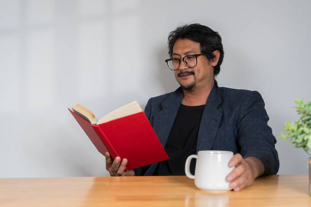 istock Mature man reading book by window, smiling wearing glasses, copy space. 2210188051