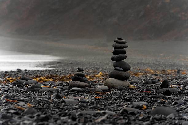 A mountain of black rocks rises above a quiet volcanic beach with dark sand under a dramatic cloudy sky