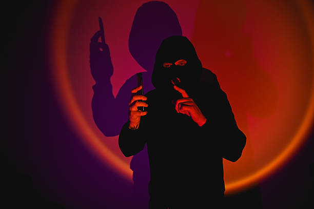 istock Masked hacker making silence gesture holding smartphone under colorful light 2210192561