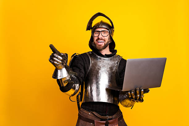 istock Young man in knight's armor with a laptop and pointing against a yellow backdrop 2210193115