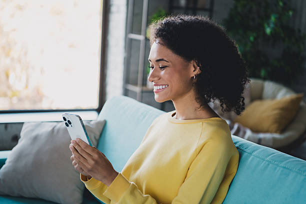 istock Smiling young woman using smartphone while sitting on comfortable sofa in a bright stylish living room interior 2210196590
