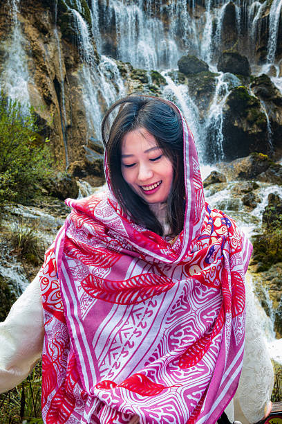 A woman stands in front of a waterfall, dressed in a white outfit with a red scarf, showing a bright smile against the stunning natural backdrop