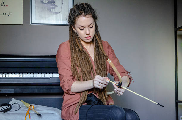 A young woman with long natural dreadlocks is crafting a bow and string in the corner of her apartment using bamboo wood and black cord. The scene reflects a blend of focus, creativity, and traditional craftsmanship in a modern indoor setting.