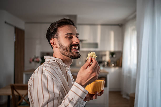 istock Happy man eating noodles in a bowl with a fork at home 2210209115