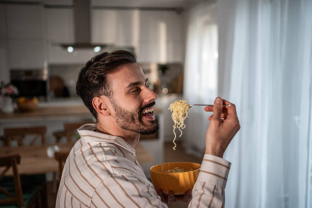 istock Happy man eating noodles in a bowl with a fork at home 2210209123
