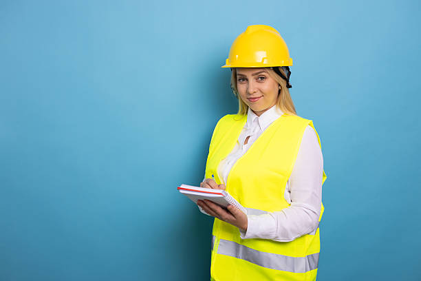 istock Confident female engineer wearing safety vest and helmet taking notes on a notepad on a blue background 2210209479
