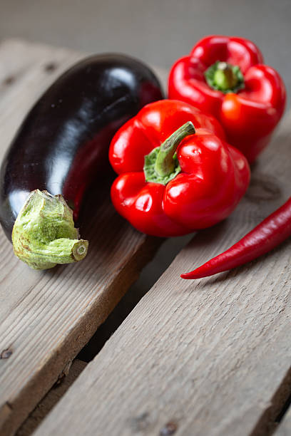Colorful fresh vegetables including eggplant, red bell peppers, and chili peppers on a weathered wooden surface. Great for Mediterranean recipes or plant-based diet visuals.