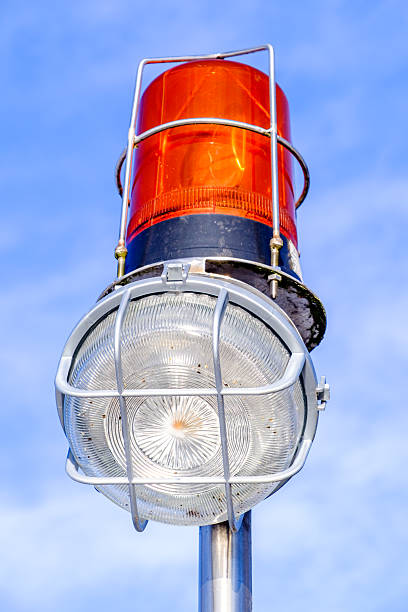 A bright orange warning light is mounted on a metallic pole with a shiny lens below it, shining vividly against a clear blue sky in daylight.