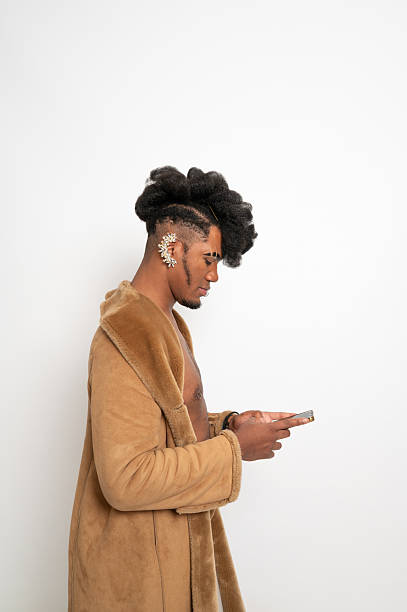 Young man wearing a fashionable fur coat and an earring, focused on using a smartphone against a clean white background