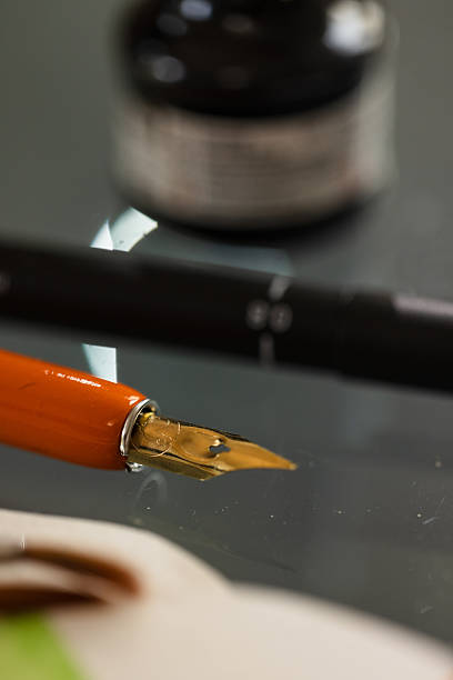 Close-up of a golden calligraphy nib with an orange handle, resting on a reflective glass surface. An ink bottle and black marker are softly blurred in the background. Ideal for artistic or vintage-themed creative visuals.