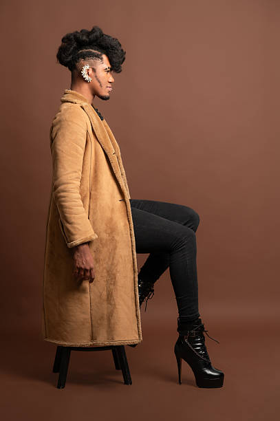 istock Stylish young man sitting on stool wearing long coat and high heels 2210269941
