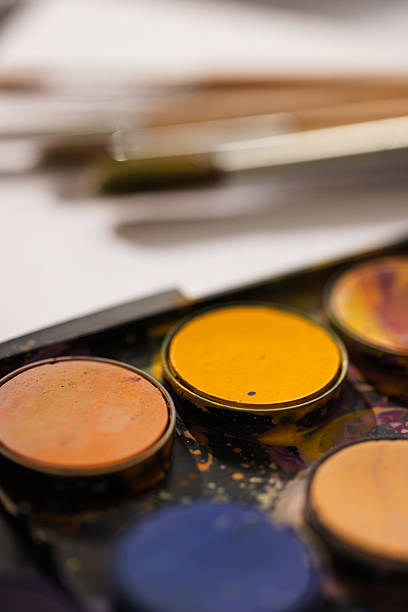 Flat lay of colorful watercolor paints and a selection of painting brushes placed on white textured paper. Clean and minimal artistic background.