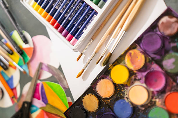 Top view of a vibrant and artistic setup featuring watercolor paints, oil pastels, brushes, scissors, colored paper cut-outs, ink, and fine liners. A perfect representation of a creative workspace, DIY projects, or school art supplies.