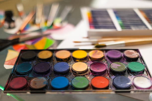 Artistic workspace featuring watercolors, oil pastels, paintbrushes, and colorful paper circles. The scene is captured with shallow depth of field to emphasize creative mood and handmade art tools.