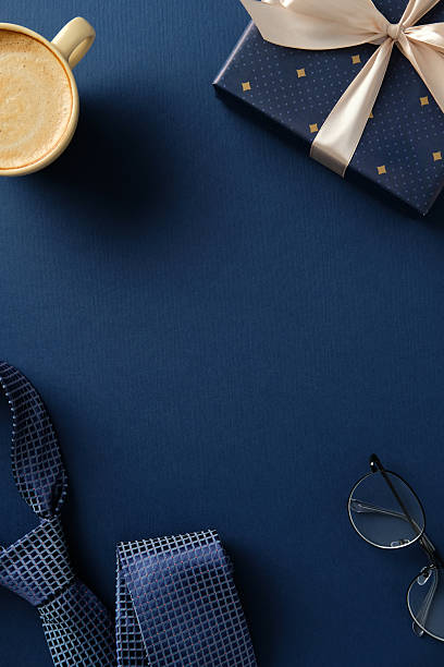 Men's accessories flat lay: Blue tie, coffee cup, gift box, and eyeglasses on a blue background. Ideal for Father's Day, business gifts, or masculine lifestyle concepts.