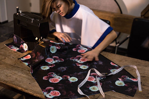 Young woman designer sewing black floral fabric on black home sewing machine on wooden table. 
Concept of hobby sewing as well as tailoring.